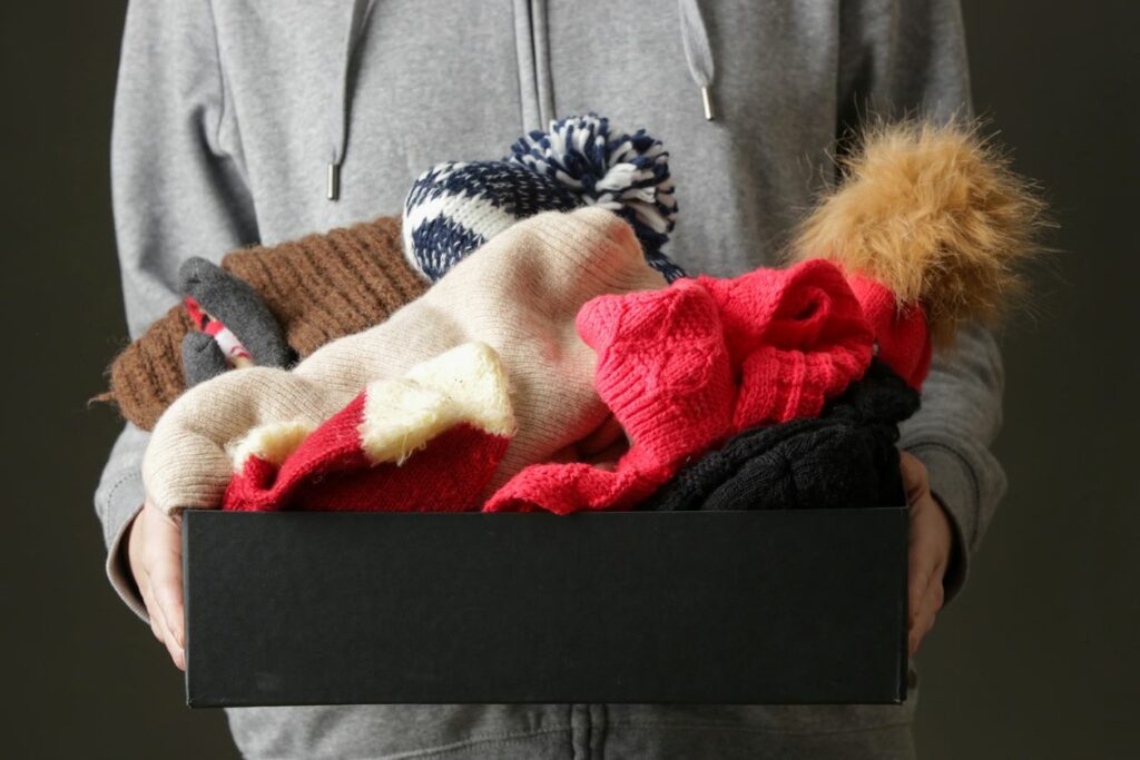 A person holds a box of winter hats, scarves, and other accessories.