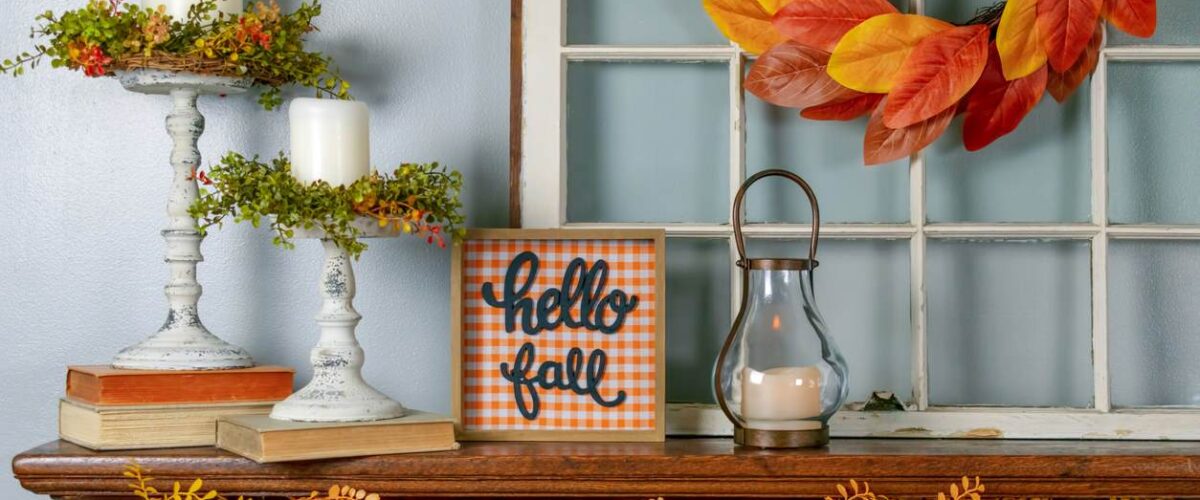 Various fall decorations, including a garland, wreath, lantern, and sign reading “Hello Fall” sit on a mantle.