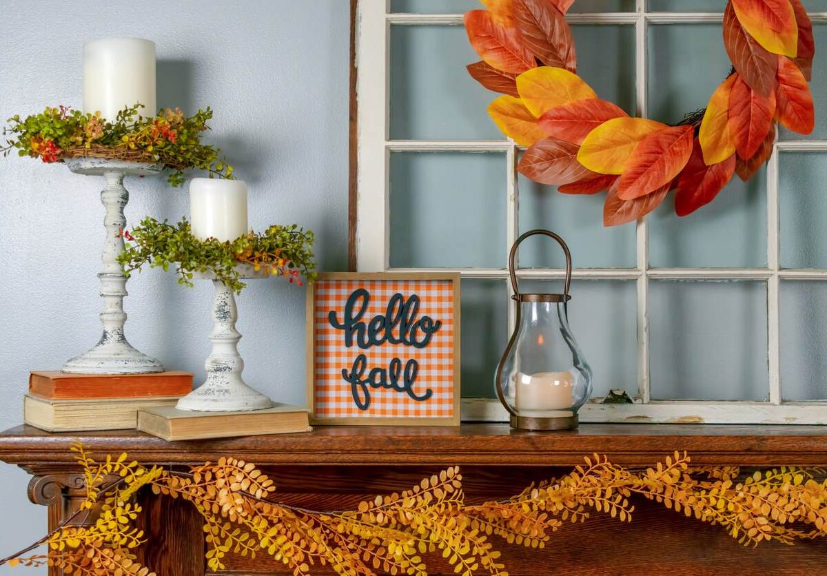 Various fall decorations, including a garland, wreath, lantern, and sign reading “Hello Fall” sit on a mantle.