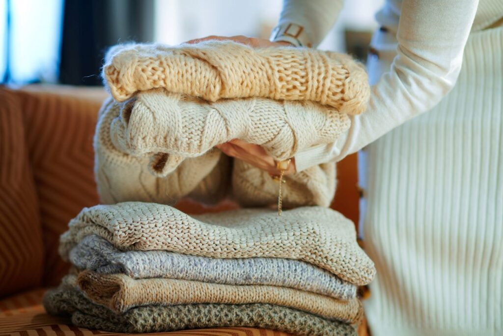 A woman lifting a stack of folded sweaters off a couch. 